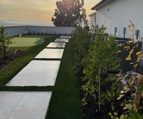 Luxury backyard landscape design with large white paver stepping stones, artificial turf, and a small putting green under a sunset sky.