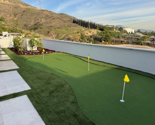 Panoramic view of a large, realistic artificial turf putting green with three yellow flags, contrasting sharply with a steep, natural brush-covered hillside.