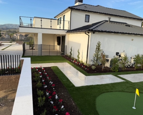Modern luxury home exterior with a black metal roof, white stucco walls, glass balcony railing, a fenced patio, and meticulously designed ground-level landscaping featuring a red and white flower bed and artificial turf.