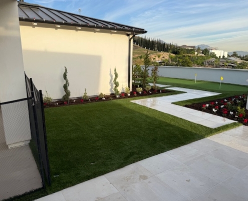 Landscape detail of a clean zigzag white paver pathway cutting through artificial turf, bordered by red and white flowers and young evergreen plantings.