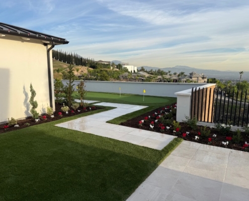Custom backyard putting green with two yellow flags, seen from a patio area. The landscape features artificial turf, white pavers, and a distant view of homes on a rolling hillside.