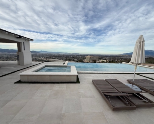 Minimalist lounge chairs and a white umbrella on a stone patio next to an infinity-edge pool.